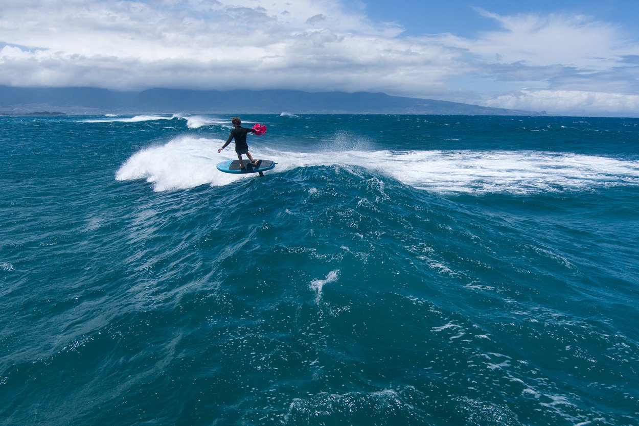 A surfer rides a blue wave in the ocean under a partly cloudy sky, with mountains and the Stash ParaWing 2025 visible in the background.
