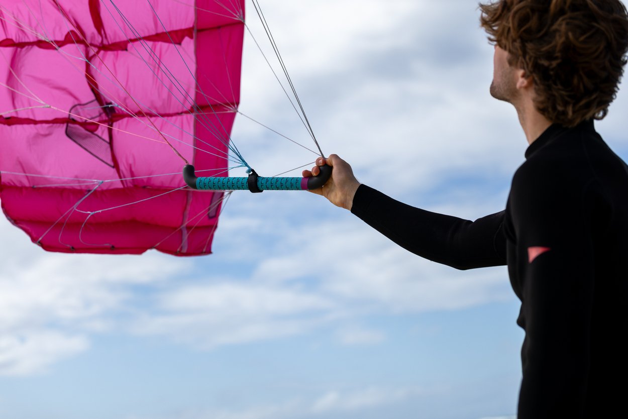 A person in a wetsuit holds the Stash ParaWing 2025 kitesurf sail, pink against a cloudy blue sky.
